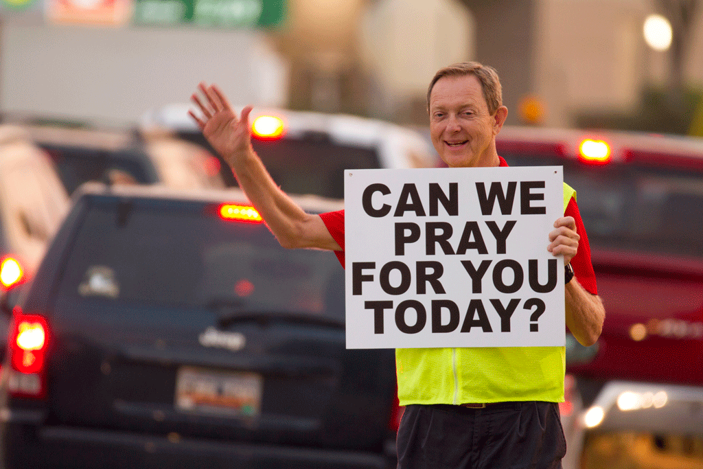 Man waving holding a can we pray for you today sign in front of cars