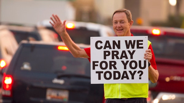 Man waving holding a can we pray for you today sign in front of cars
