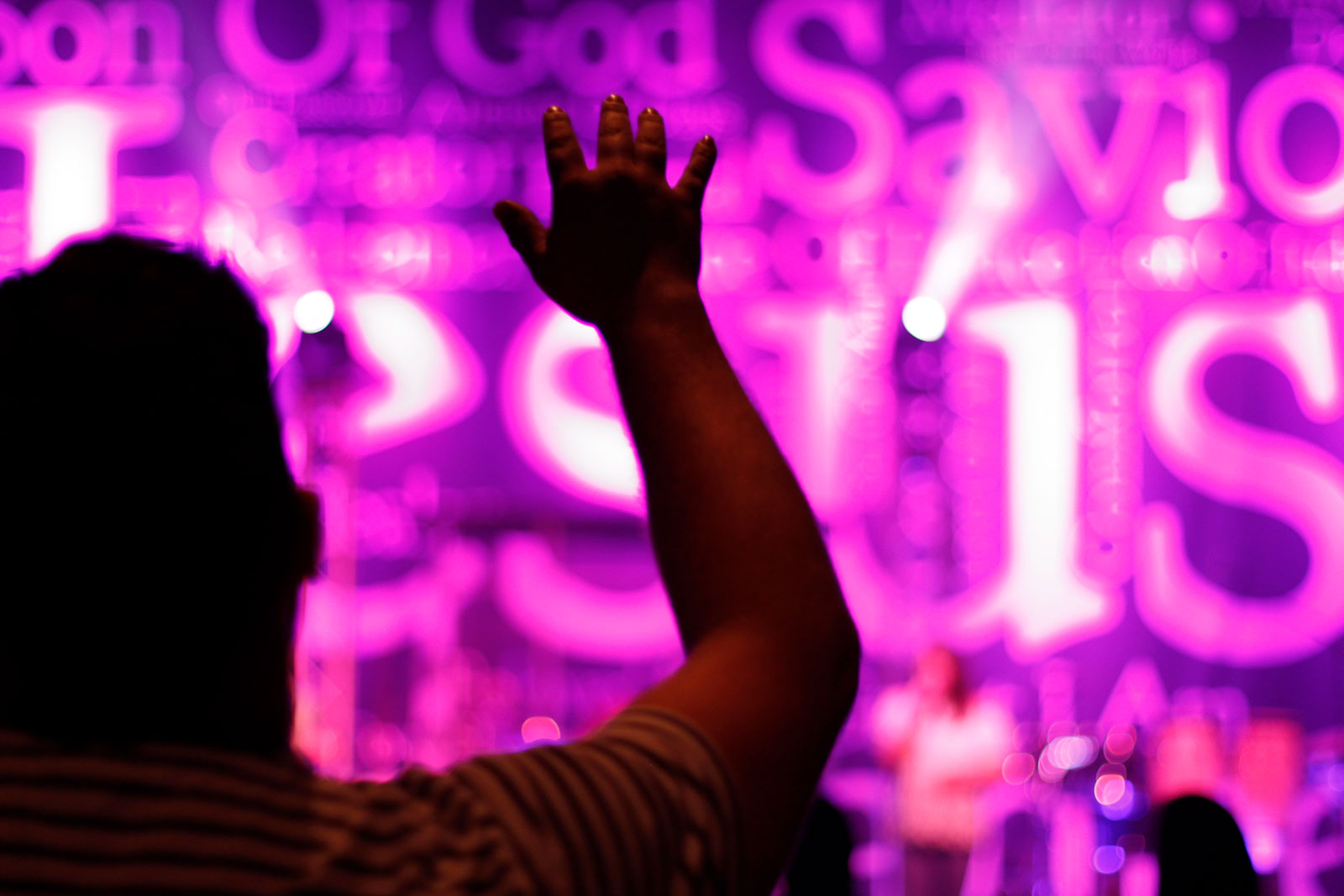 Person holding up hand worshipping in front of a purple lit Jesus banner during Contemporary Services at Saxe Gotha